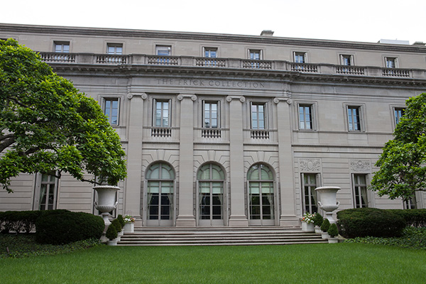 The stone facade of the Frick Collection. A stairway goes from a grass lawn up to three arched glass doors.