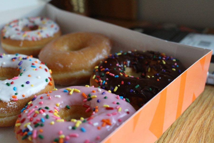 Five doughnuts from Dunkin’ rest in an orange box on a light brown wooden table. Out of the four doughnuts, two are glazed with white icing and sprinkles, one is glazed with pink frosting and sprinkles, one is glazed with brown frosting and sprinkles, and the last one is plain glazed. A book can be seen in the distant background towards the right corner of the image.