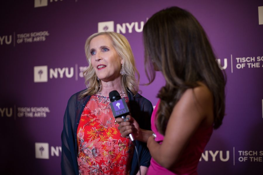 A blonde woman speaks into an N.Y.U microphone in front of a purple N.Y.U. backdrop.