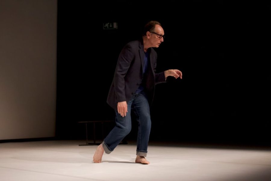 Paul Lazar walks barefoot across a stage. He is wearing a black blazer, royal blue T-shirt and denim jeans that are cuffed at the ankle.