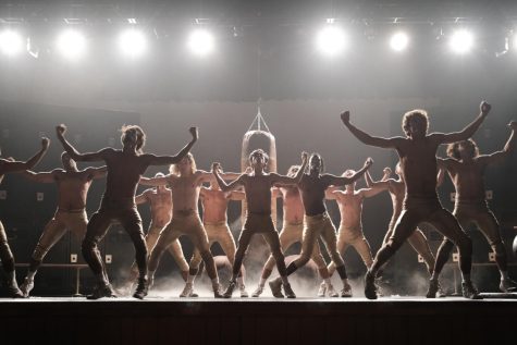 Thirteen high school boys perform a dance onstage while wearing football pants. Behind them are prop lockers and a punching bag that hangs from the center of the ceiling.