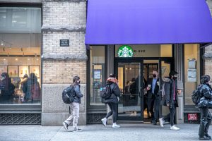 Facade of the Starbucks coffee shop at NYU’s Goddard Hall. Some pedestrians are walking in front of the building and a customer is opening the front door.
