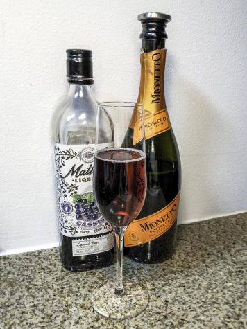 Two alcohol bottles and a champagne flute on a granite countertop. On the left, a mostly empty bottle of liqueur. On the right, a half-empty bottle of prosecco with an orange decal. In the center, a champagne flute with a homemade deep red spritz.