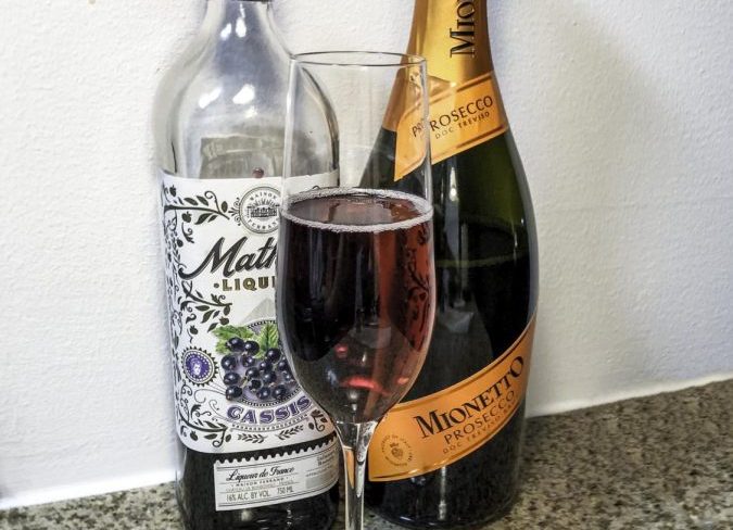 Two alcohol bottles and a champagne flute on a granite countertop. On the left, a mostly empty bottle of liqueur. On the right, a half-empty bottle of prosecco with an orange decal. In the center, a champagne flute with a homemade deep red spritz.
