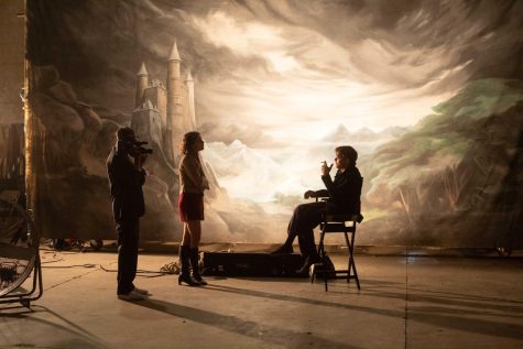 On the left, Marland props a video camera on his shoulder. In the center, Garance stands with her shoulder crossed. On the right, Jim sits in a directors chair while smoking a cigarette. All three individuals are on a film set in front of a painted tarp that depicts a castle in the midst of mountainous terrain.