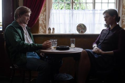 Max and Julie sit at a cafe table next to the window. Max is wearing a green zip jacket, white collared shirt, and blue jeans. Julie is wearing a navy blue knee length skirt, a grape collared shirt, and a deeper purple fully buttoned vest.