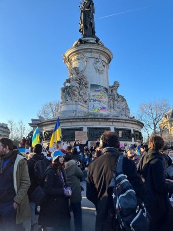 A group of people congregate around a monument with homemade banners. Some hold Ukrainian flags, while others hold cardboard signs with messages written in black ink.