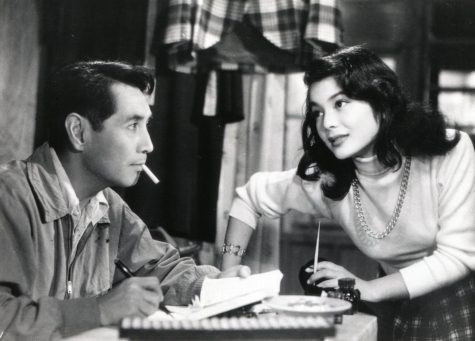 A black-and-white picture of actor Masayuki Mori sitting with a cigarette in his mouth and a pen in hand, writing in a notebook. In front of him, actress Yoshiki Kuga looks at him, standing and with her hands on the table.