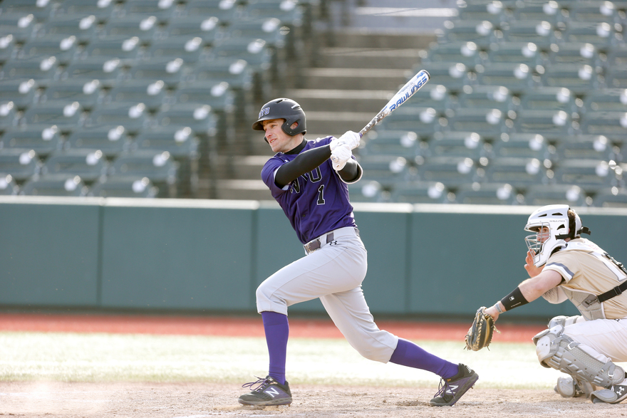 NYU baseball player Grant Berman follows through on his swing after hitting a pitch. He wears a purple jersey and white pants cuffed at the knee to reveal purple socks. On the right, a catcher crouches on the ground in an all-white uniform.