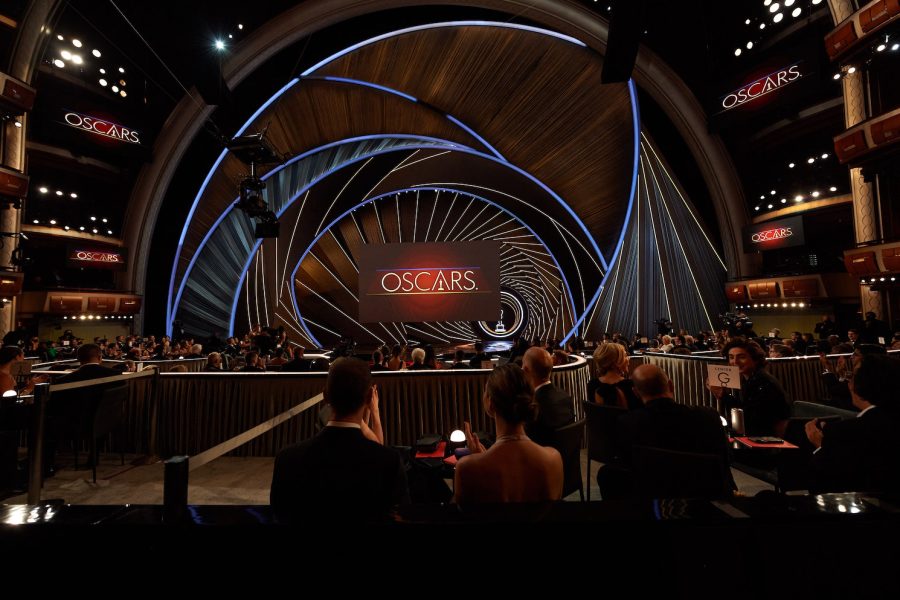 A wide-angle photograph of the Dolby Theater in Los Angeles, where the 2022 Academy Awards were held. Hundreds of guests fill the auditorium. In the foreground, two guests sit at a table wearing red carpet attire. In the background, the Oscars logo is shown on a screen mounted onto the stage.