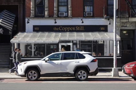 The facade and entrance of The Copper Still. The building and patio awning are white and have black trim around the windows and doors. Above the entrance are the words “The Copper Still” in gold lettering.
