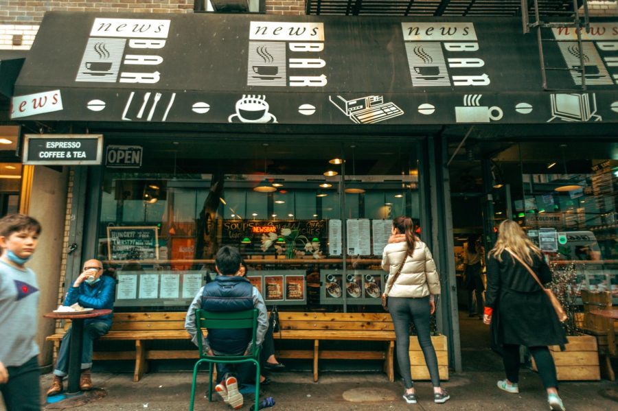 The storefront of Newsbar at 107 University Place. The door is open and people are seated outside. Others walk past the cafe.