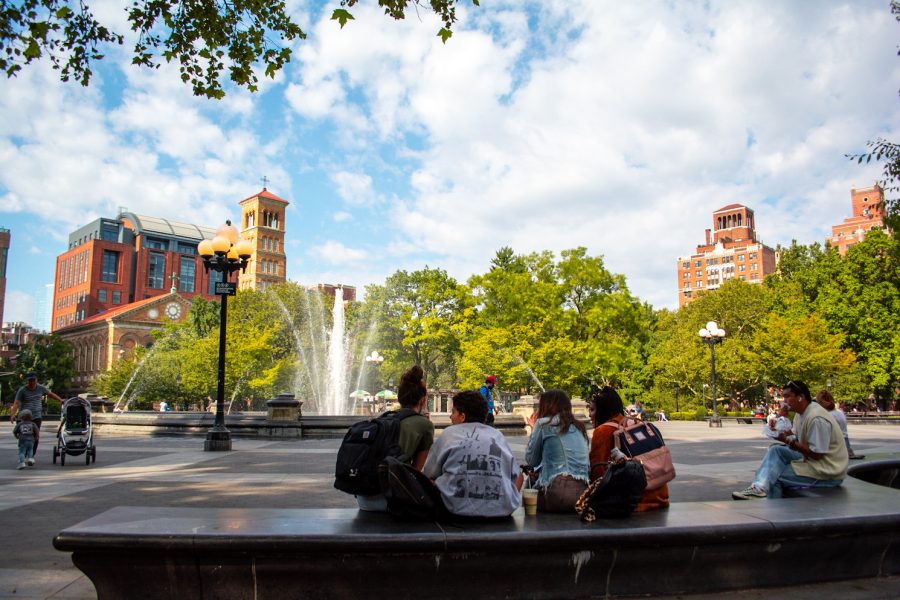 A group of students sit on a bench in Washington Square Park. Buildings in the background rise above green trees with lots of foliage. It is a sunny day with a bright blue sky full of clouds. The fountain in the center of the park is on and spewing water into the air.