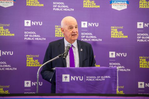 NYU president Andrew Hamilton delivering a speech at a podium. On the podium, a purple banner hangs with the NYU logo and the words “School of Global Public Health” in white letters. Hamilton is wearing a black blazer, white button-up shirt and bright purple tie.