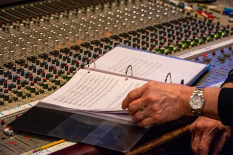 An open binder full of sheet music sitting on top of a sound board. A hand wearing a silver watch holds one of the pages in the binder.