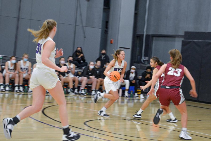 Belle Pellecchia dribbles a basketball across the court toward the basket. She is wearing an all-white uniform with her hair pulled back in a ponytail. Around her is one of her teammates and two players from the opposing team