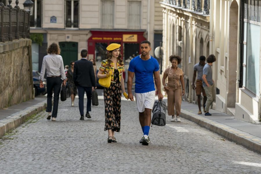 A still from the television show “Emily in Paris,” in which characters Emily and Alfie walk down a Parisian street holding hands. Emily is wearing a black dress with a yellow cardigan, bag, beret, and gloves. Alfie is wearing white athletic shorts and a blue jersey.