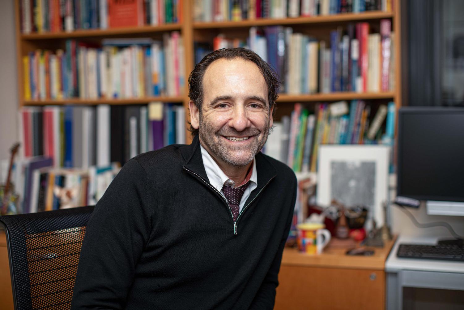 A portrait of Dr. Jess Shatkin in his office. He is sitting in a desk chair in front of a crowded bookshelf. He is wearing a black quarter-zip sweatshirt, a white collared shirt and a maroon tie.