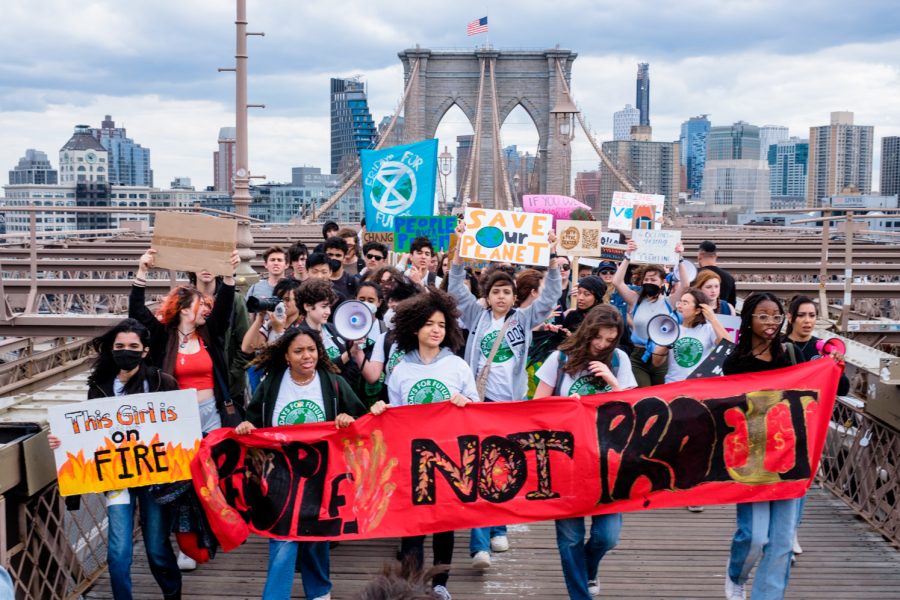 A crowd of mainly young protestors hold up a red banner saying "People Not Profit" and other protest signs on a march across the Brooklyn Bridge pedestrian path on a cloudy day as an American flag waves in the wind on top of the bridge's arches.