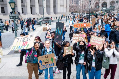 A group of protesters hold up signs on the sidewalk of Foley Square as pedestrians and drivers pass by on the nearby street.