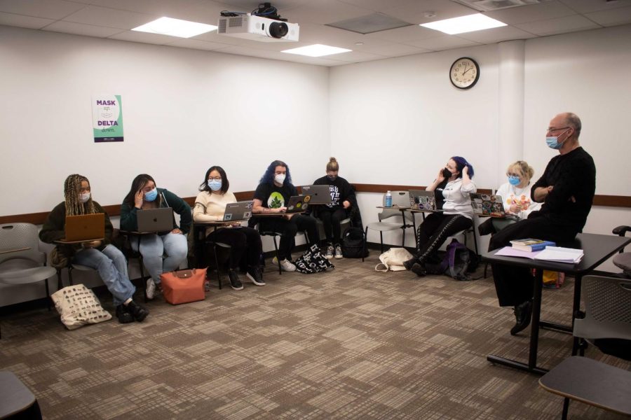Seven students wear masks while sitting at their desks in an NYU classroom. A professor sitting on a table listens to a student.