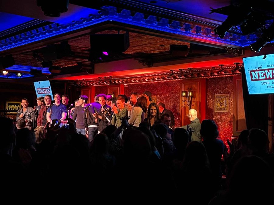 Multiple “Newsies” cast members stand behind microphones on a small stage surrounded by an applauding crowd.