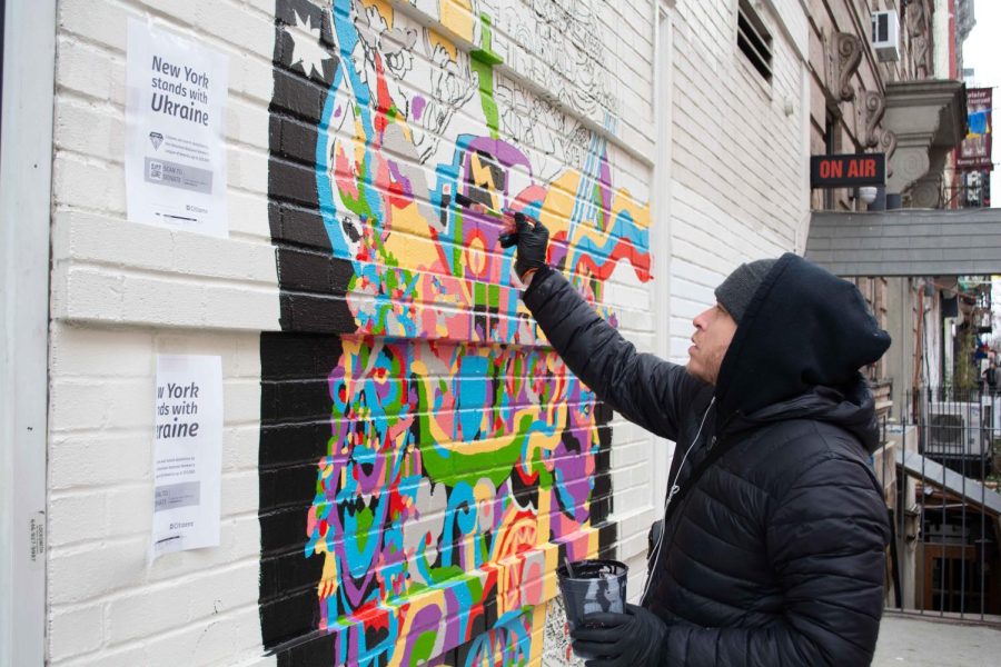 Misha+Tyutyunik+painting+a+colorful+mural+on+a+beige+brick+wall.+He+is+wearing+a+black+jacket%2C+hoodie+and+beanie.+He+holds+a+paintbrush+in+his+right+hand+and+a+cup+of+black+paint+in+his+left.