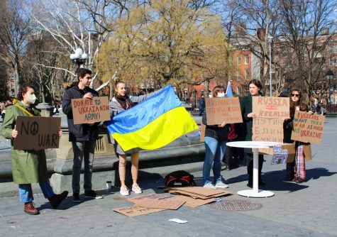 Six protesters stand by the Washington Square Park fountain holding up cardboard posters and a Ukrainian flag in support of ending the invasion of Ukraine by Russia.