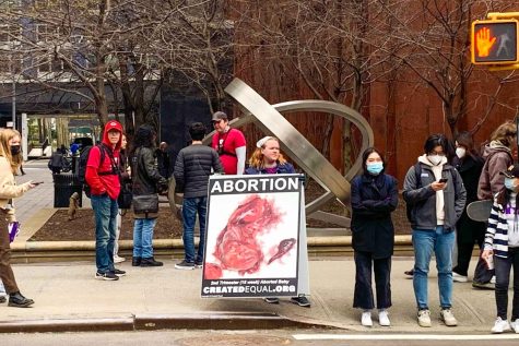 A woman stands on the sidewalk in front of NYU’s Bobst Library holding a big sign that shows a graphic image of a fetus, and above it the word “Abortion.” Next to the woman, students with backpacks head to class or are waiting to cross the street.