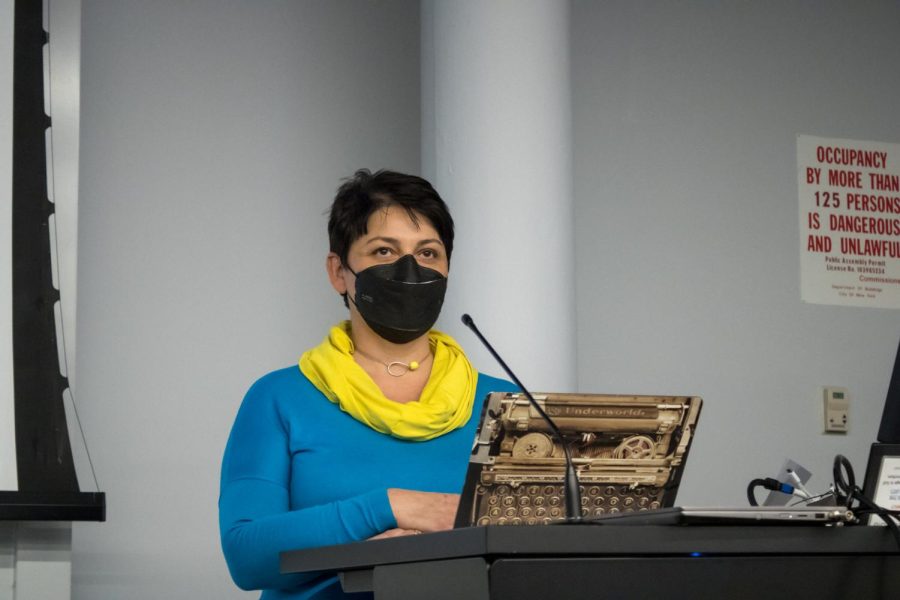 Natalia Levina stands behind a podium and addresses the crowd. She is wearing a bright blue long-sleeve shirt and a yellow wrap scarf.