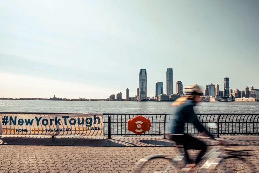 A blurred biker rides along the Hudson River Park piers. On the left, a white banner hangs on a rail with the words “#NewYorkTough” in royal blue. Behind both is the Jersey City skyline.