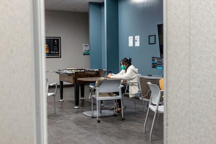 A masked student sits in an NYU common room area. She is surrounded by tables, chairs and a foosball table.