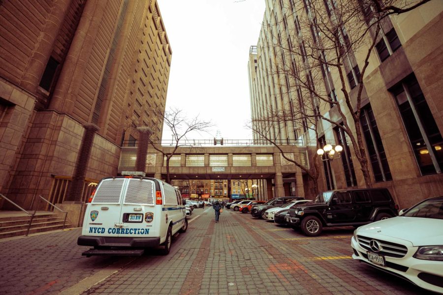 The exterior of the Manhattan Detention Complex. A skyway bridge connects both sides of the brown building. On the ground below, cars are parked in parking spots marked with yellow lines on the right side, and a van with the New York Police Department logo and the text “NYCD Correction” on the back is parked on the left side.