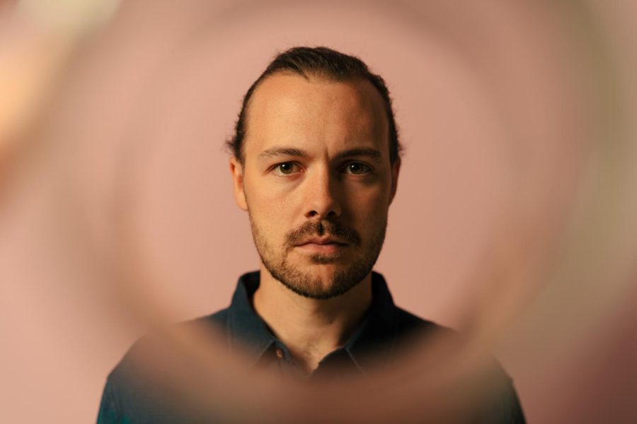 A focused headshot of Sam Webber in front of a pink background with a blurred ring circling around his head and brighter lighting focused on the left side of his face.
