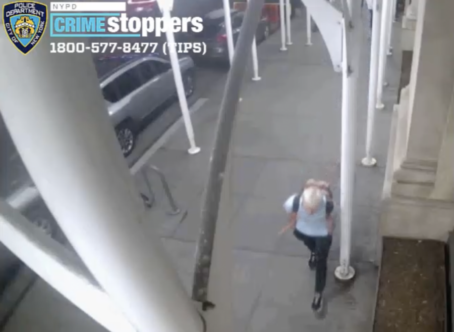 A screenshot from CCTV video released by the NYPD Crime Stoppers Program. From above, a white-haired individual wearing a light blue short-sleeve T-shirt, dark-colored slim pants walks under scaffolding.