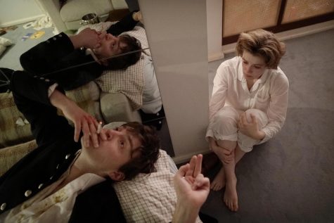 Julie sits on the bedroom floor wearing white pajamas. To her left, Anthony lies on the bed and smokes a cigarette. Next to him, there’s a mirror that reflects his image.
