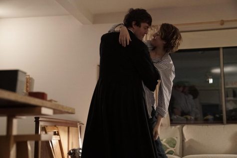 Julie and Anthony share an embrace in the middle of the living room. Anthony wears a black overcoat while Julie wears a blue and white pinstripe collared shirt and blue jeans.