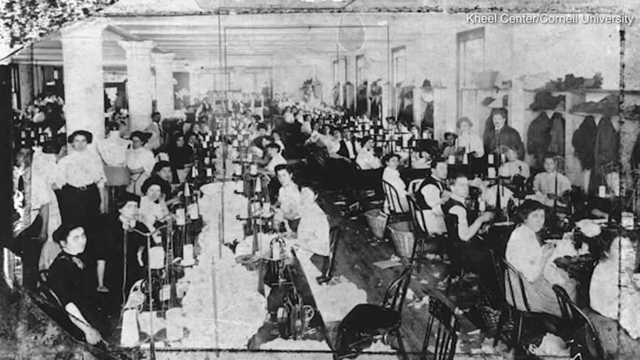 A black-and-white image showcasing the Triangle Shirtwaist Factory located in what is now the Brown Building. Two rows of factory workers sit in a long-desk formation working at sewing machines.