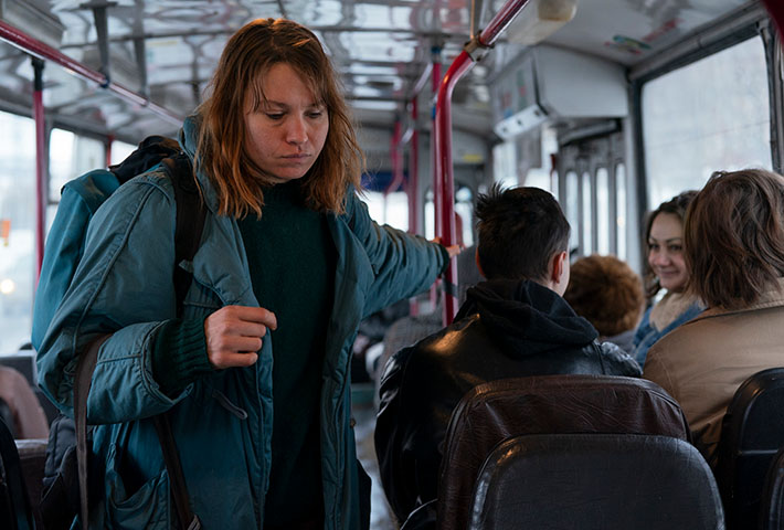 A woman with a tired face wearing a dark teal jacket and a black backpack stands and holds onto a red pole of a bus shuttle as smiling passengers are sitting down.