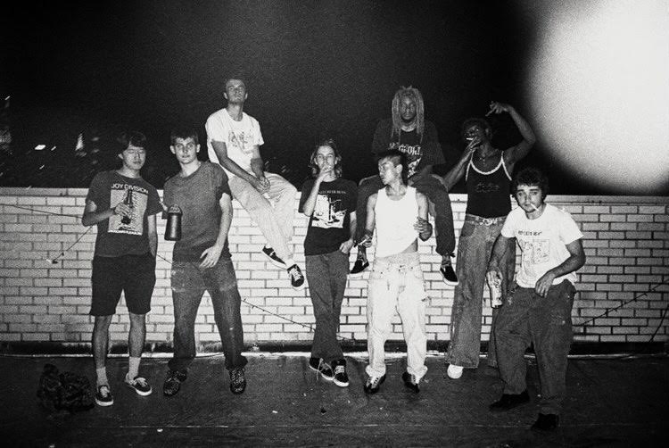 A+black-and-white+group+portrait+shot+on+film+of+six+skateboarders+on+a+rooftop.+Two+members+of+the+group+are+sitting+on+the+wall%2C+white+the+rest+stand+against+it.+A+few+members+of+the+group+hold+cigarettes+or+bottles.
