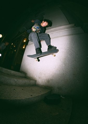 Yoshi performs a trick on his skateboard as he rides over a staircase at night. He is wearing a black graphic short-sleeve shirt and black denim pants.