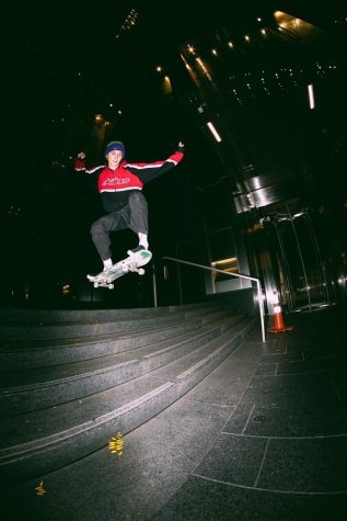 Simon performs a trick on his skateboard as he rides over a staircase at night. He is wearing a red-and-blue zip-up hoodie with black pants cuffed at the ankle.