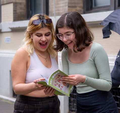 A portrait of Natalia Palacino and Susan Behrends Valenzuela reading a zine. Palacino is wearing a white scoop neck tank while Behrends Valenzuela is wearing a mint-green long-sleeve shirt and a black skirt.