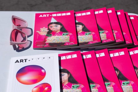 An assortment of hot pink zines that read “Art + Type” on a white card table. On the left, there’s a pair of square pink sunglasses.