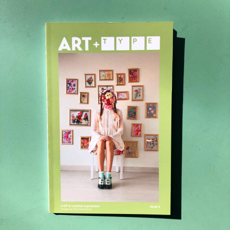 A lime-green zine that reads “Art + Type” on a deeper forest green background. On the front of the zine is a photograph of a person sitting in front of a wall of paintings with a bouquet of flowers obscuring her face.