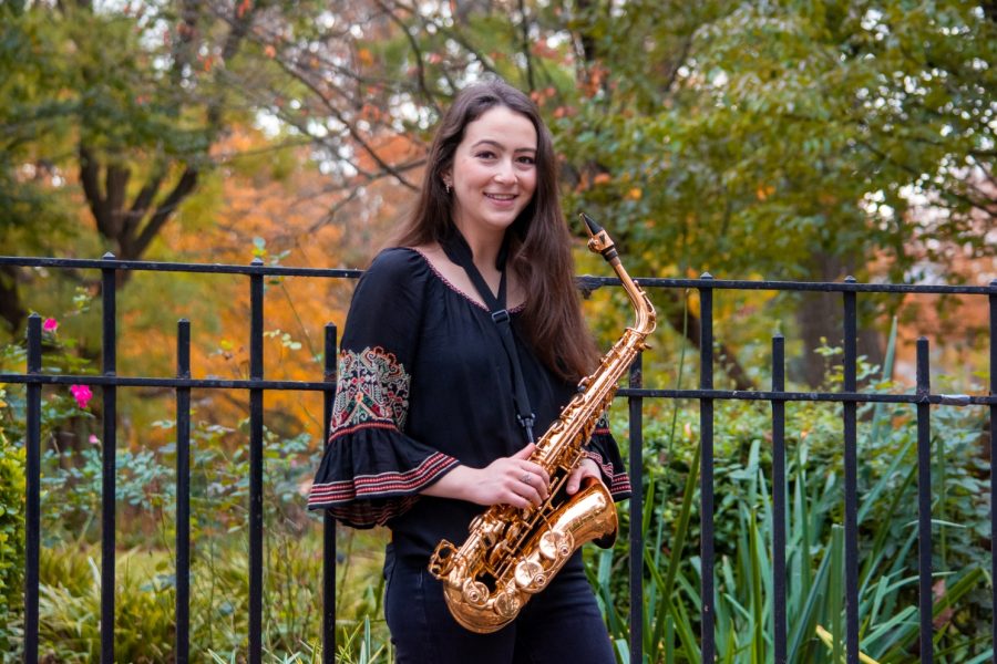 A+portrait+of+Olivia+Hughart+in+Tompkins+Square+Park.+She+is+wearing+a+navy+blue+three-quarter+sleeve+blouse+with+a+ruffle+hem+at+the+elbow.+She+is+holding+a+saxophone+in+her+hands.