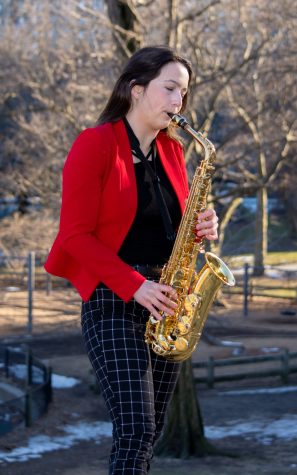 Olivia Hughart playing a saxophone in Central Park. She is wearing a red blazer over a black blouse, and black-and-white geometrically patterned pants.