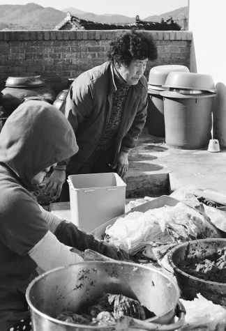 A black-and-white photo of Asian men cooking outdoors over large pots. Behind them is a brick wall and a mountainous landscape.