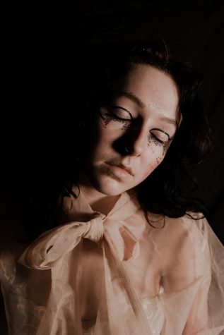 A studio portrait of a girl with her eyes closed. She is wearing a cream sheer blouse that ties in a bow at the neck. She is also wearing thick black eyeliner and glitter across her cheeks.