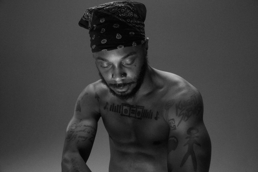 The musician JPEGMAFIA wearing a bandana. He has a beard and tattoos on his chest and arms. He has his eyes closed and is looking down.